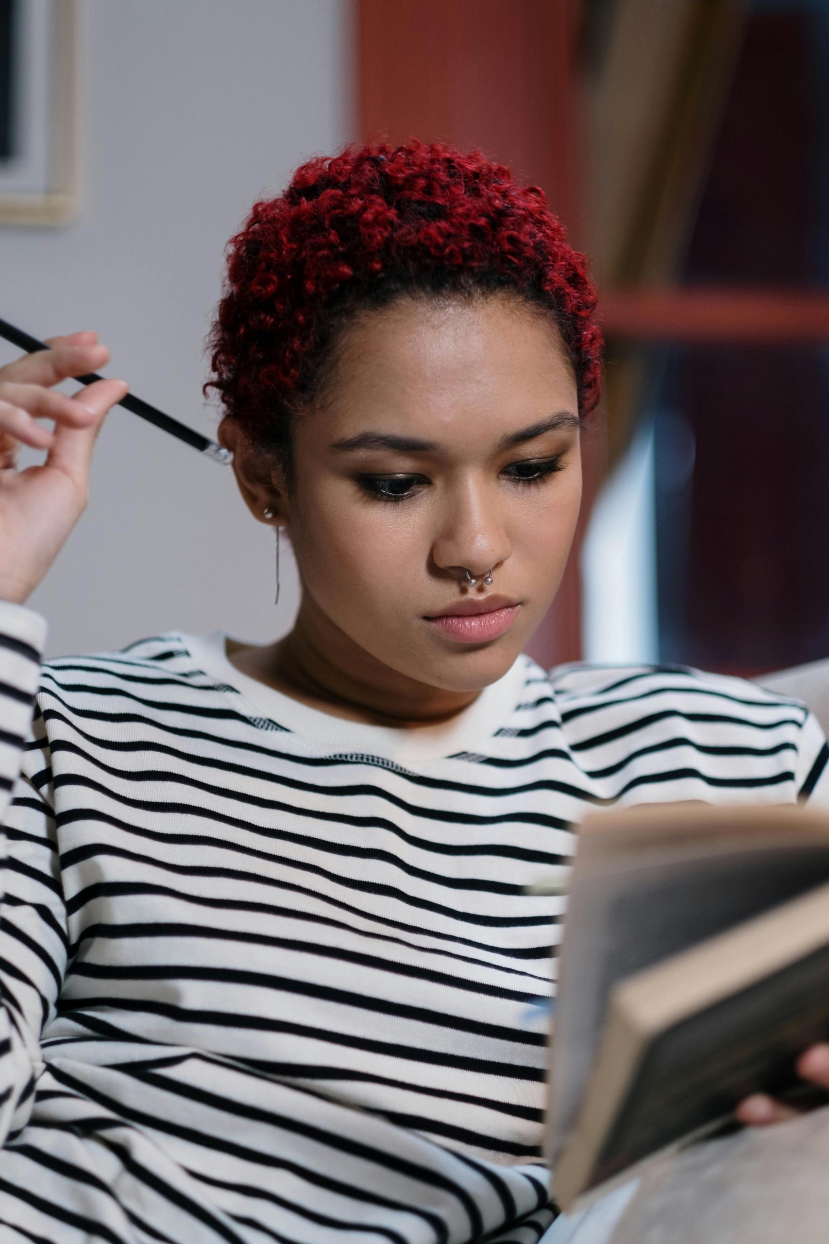 A young woman with curly red hair and nose piercing focused on studying indoors.